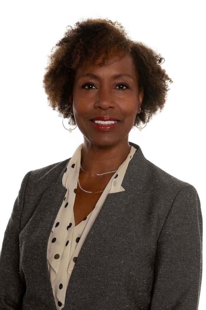 Lynne usa blazer cinza e está de pé, sorrindo. 