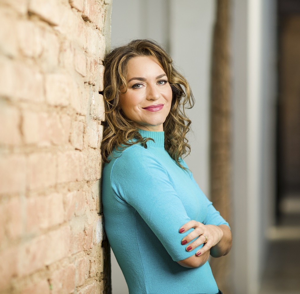 Luciana está encostada na parede, de braços cruzados, olha para frente e sorri